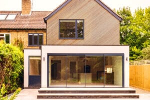 Modern two-story house featuring aluminium casement windows and a glass door extension, set against a backdrop of greenery and a wooden fence.