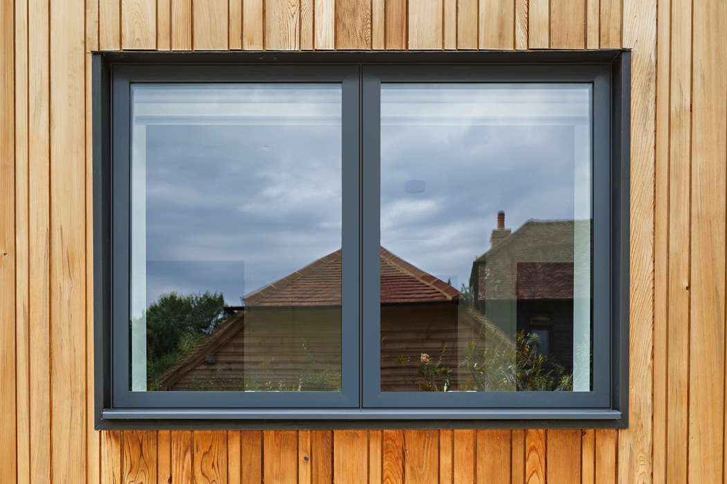 An aluminium casement window with a gray frame is set against a wooden wall. The reflection captures buildings, plants, and a cloudy sky.