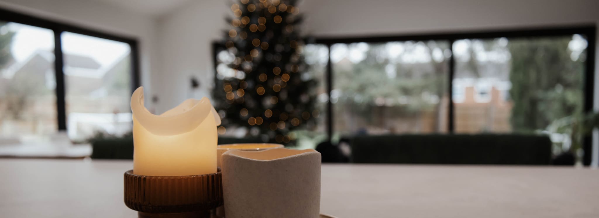 Two flameless candles sit on a table indoors, with a blurred, lit Christmas tree and large windows in the background.