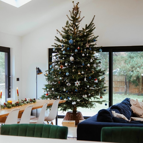 A large Christmas tree with various ornaments stands in a bright, modern living room. Bifold doors open to reveal a dining table elegantly decorated with candles and greenery, creating a seamless connection between indoor warmth and outdoor beauty.