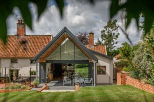 A modern house with a glass-fronted extension and gable window features an outdoor dining area and a red tiled roof, all set in a garden adorned with trees and encircled by a brick wall.