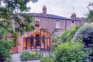 A modern glass and wood extension at the back of a brick townhouse features a striking gable window, surrounded by lush greenery and a garden path.