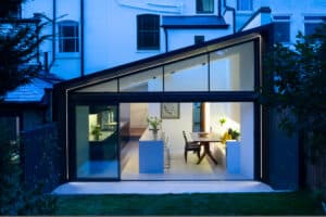 Modern house with a large glass extension and gable window, featuring a lit interior with a kitchen island and dining area, surrounded by garden greenery.