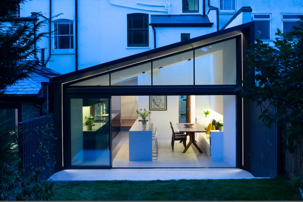 Modern house with a large glass extension and gable window, featuring a lit interior with a kitchen island and dining area, surrounded by garden greenery.