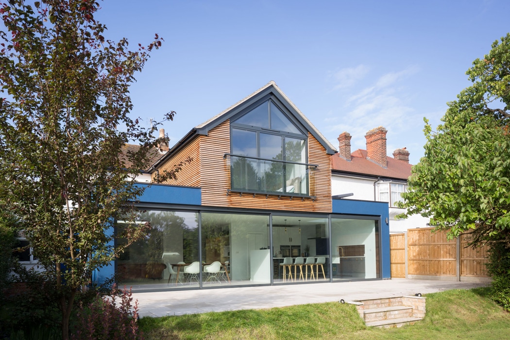 A modern house with large glass doors and a wooden facade, featuring a gable window. The spacious patio and lawn in the foreground are surrounded by trees and neighboring houses.