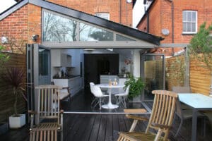 A modern extension of a brick house features large open glass doors, revealing a kitchen and dining area with a round table and chairs beneath a stylish gable window. The wooden outdoor deck is adorned with patio furniture and lush plants, creating an inviting space for relaxation.