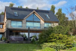 A modern, two-story house features expansive glass windows and a manicured lawn. A gable window accentuates the dark roof, with trees elegantly framing the background.