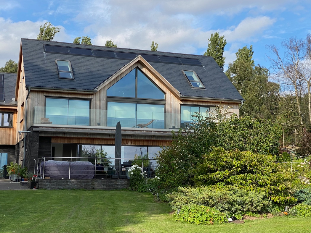 A modern, two-story house features expansive glass windows and a manicured lawn. A gable window accentuates the dark roof, with trees elegantly framing the background.