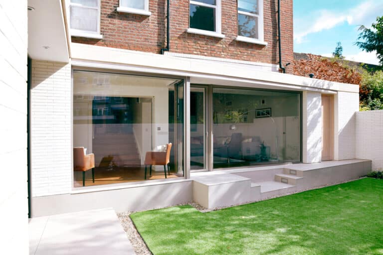 A modern house extension with large glass windows, brick exterior, and a neatly trimmed grass lawn. Two armchairs are visible through the windows.