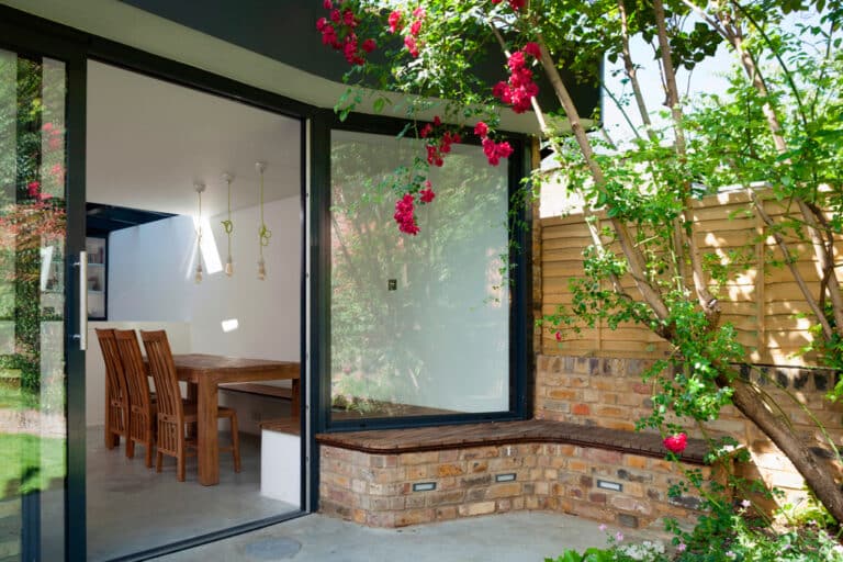 Modern dining area visible through large glass door, with wooden chairs and table. Outside, a brick wall with a built-in seat and flowering tree next to a wooden fence.