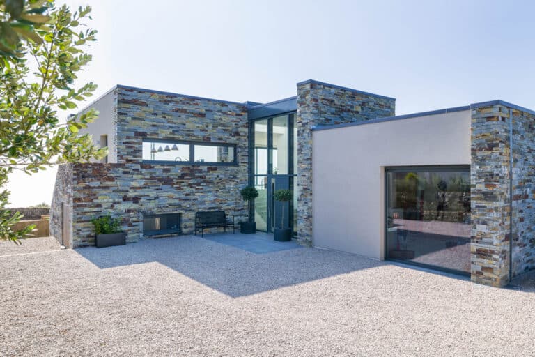 Modern house with stone and stucco exterior, large windows, and gravel driveway. Bushes are visible on the left.