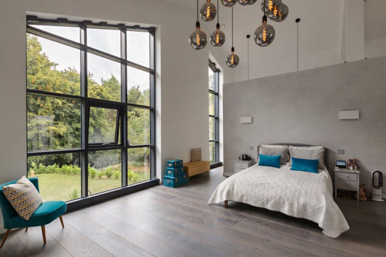 Modern bedroom with large windows, a double bed with blue pillows, and wooden flooring. There are luggage bags stacked by the window and a turquoise chair beside a small cushion.