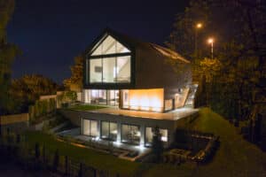 Modern, multi-story house with large glass windows and illuminated interiors, set on a landscaped slope at night.