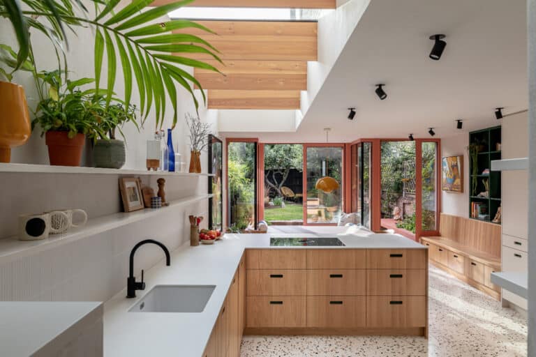 Modern kitchen with skylights, wooden cabinets, and a terrazzo floor. Large windows overlook a garden, and a central island with a sink and faucet is present. Plants and decor items are on shelves.