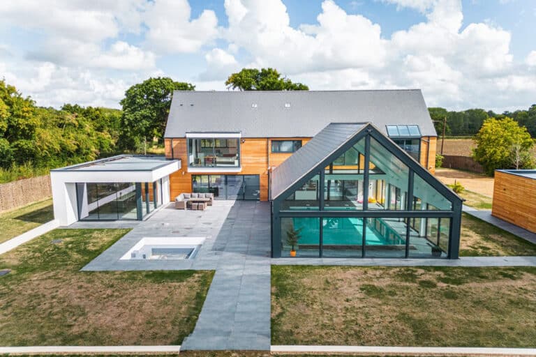 Modern house with a glass-walled extension featuring an indoor pool, surrounded by a patio and grassy lawn under a partly cloudy sky.