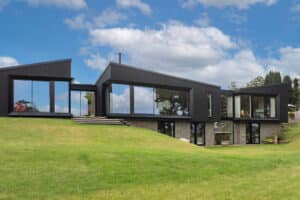 Modern black house with large glass windows on a green grassy hill under a blue sky with clouds.