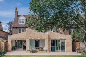 A modern brick extension with large glass doors is attached to a traditional two-story house, set in a garden with trees and greenery.