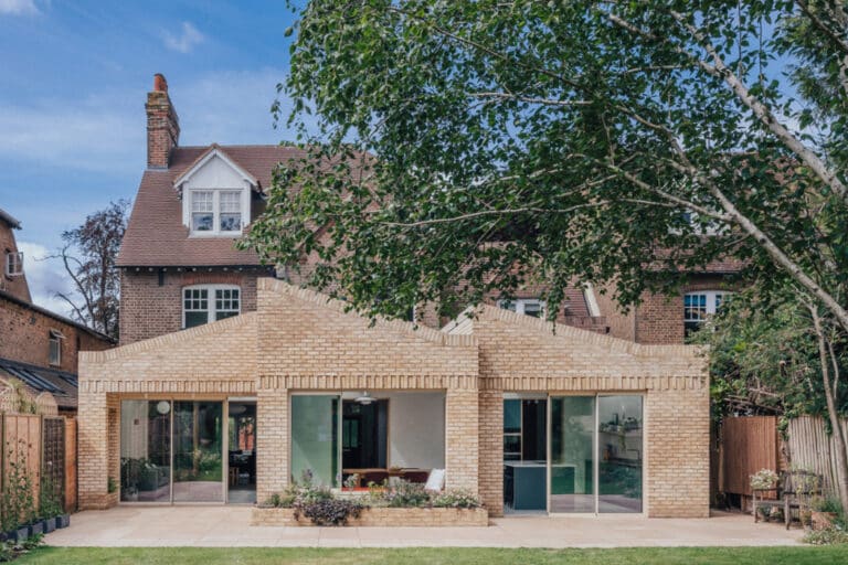 A modern brick extension with large glass doors is attached to a traditional two-story house, set in a garden with trees and greenery.