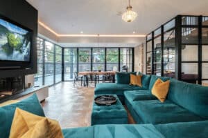 Spacious modern living room with a teal sectional sofa, yellow pillows, large windows, a TV, and a chandelier. Glass partition separates the dining area with a long wooden table.