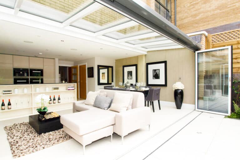 Modern open-plan living area with a beige sofa, glass wall doors, dining table, and kitchen. Walls are adorned with framed art. White and neutral tones dominate the decor.