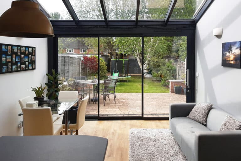 A modern conservatory with glass walls and ceiling, featuring a dining table and chairs. Outside, there is a brick patio with garden furniture and a green garden with trees.