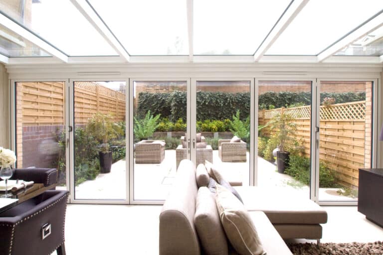 A modern living room with beige sofas opens to a patio through glass sliding doors. The patio features wicker furniture and is surrounded by wooden fencing and greenery.