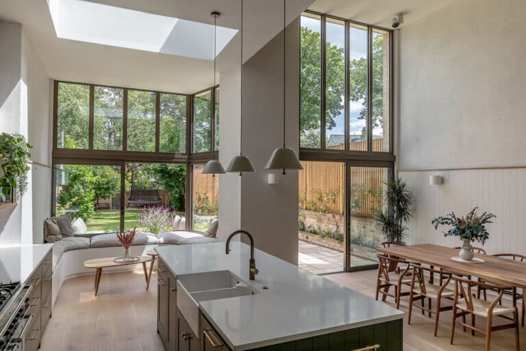 Modern kitchen and dining area with large windows, skylight, and views of a green garden. Features an island with a sink, pendant lights, a wooden table with chairs, and a cushioned seating area.