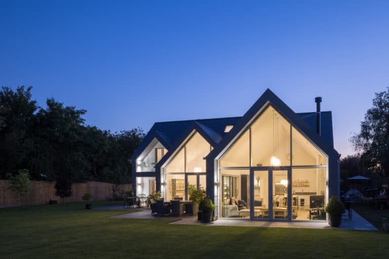 Modern house with large glass windows and sloped roof, illuminated at dusk. Green lawn surrounds the house, with trees in the background.