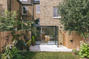 A modern brick house with a glass extension in the backyard features potted plants, a small garden, and a wooden fence.