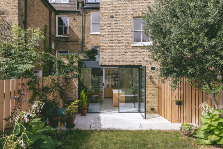 A modern brick house with a glass extension in the backyard features potted plants, a small garden, and a wooden fence.