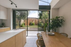 Modern kitchen and dining area with wooden furniture, large glass doors opening to a fenced garden, and potted plants.