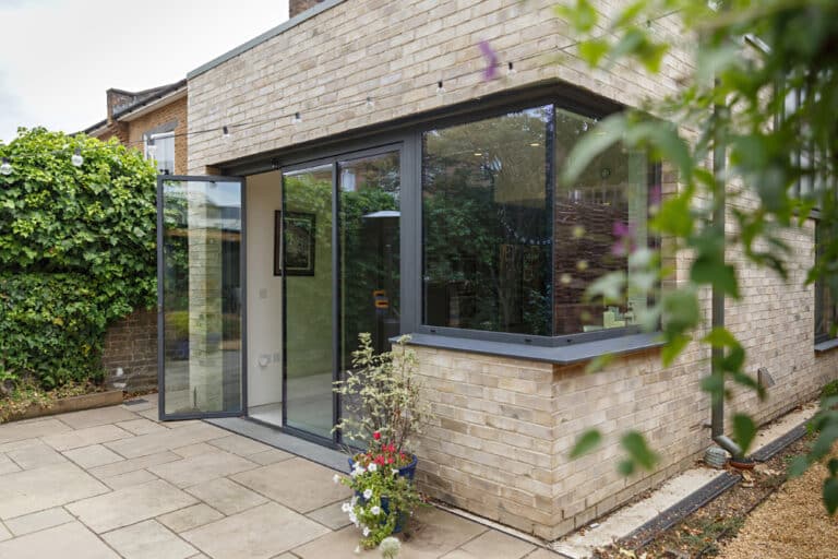 Modern house extension with brick walls, large glass doors, and a potted plant on a paved patio. Lush green foliage surrounds the area.