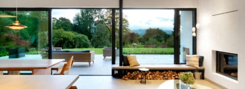 Modern living room with large glass doors opening to a patio and garden. Minimalist decor includes a fireplace, wooden bench with cushions, and contemporary lighting.