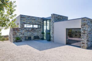 Modern house with stone and stucco exterior, featuring large gable windows and a gravel driveway, surrounded by minimal greenery.