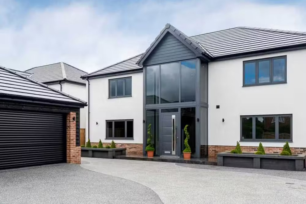 Modern two-story house with a large glass entrance, white facade, and dark gray roof featuring a sleek gable window. Double garage on the left and potted plants near the entrance enhance the home's curb appeal under an overcast sky.