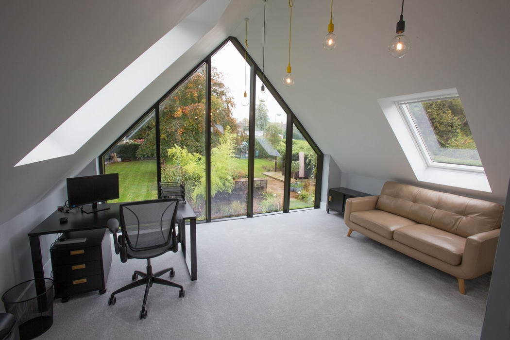 Modern attic office featuring a sleek gable window as the triangular glass wall. Inside, a desk with computer, swivel chair, and tan sofa complement the serene garden view. Three exposed light bulbs hang from the ceiling, adding an industrial touch.