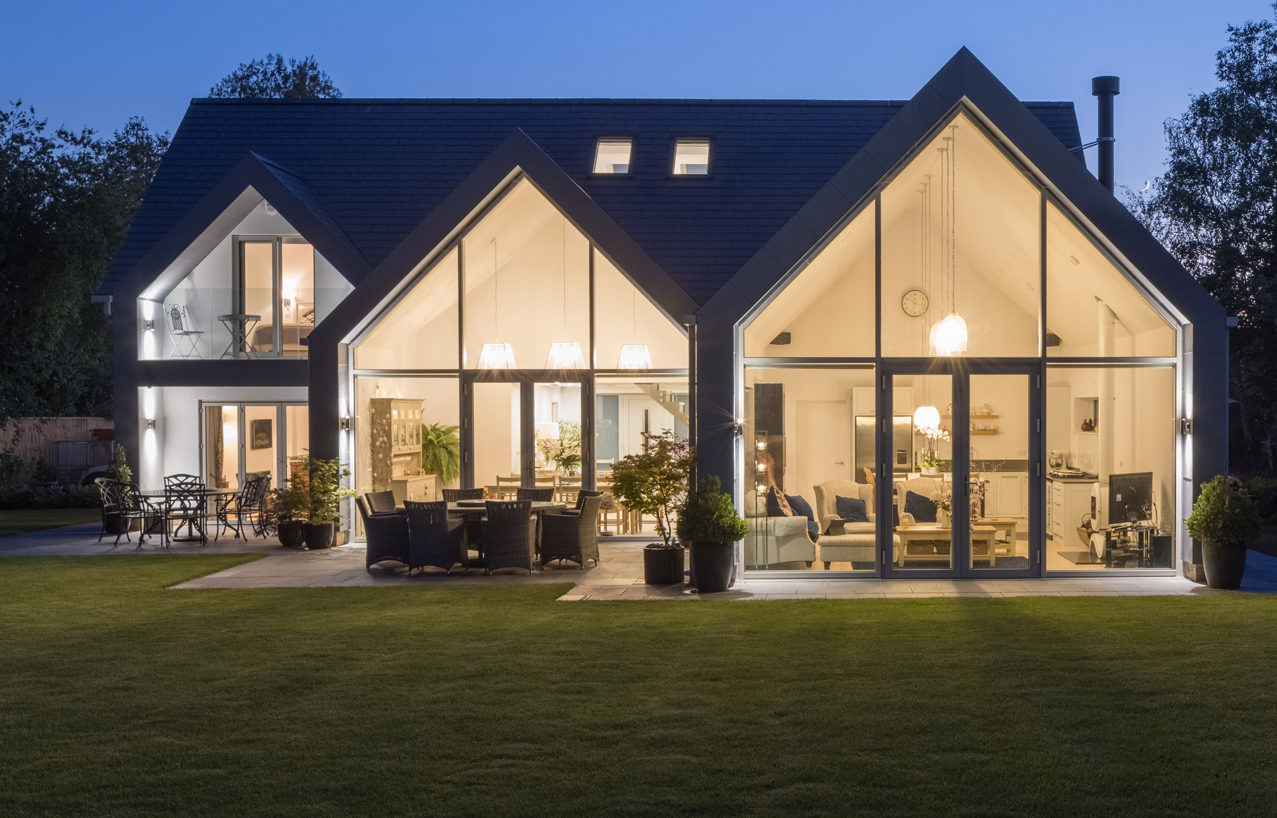 Modern house with large glass and gable windows illuminated from within at dusk, showcasing an open-plan interior and outdoor patio furniture on a neatly manicured lawn.