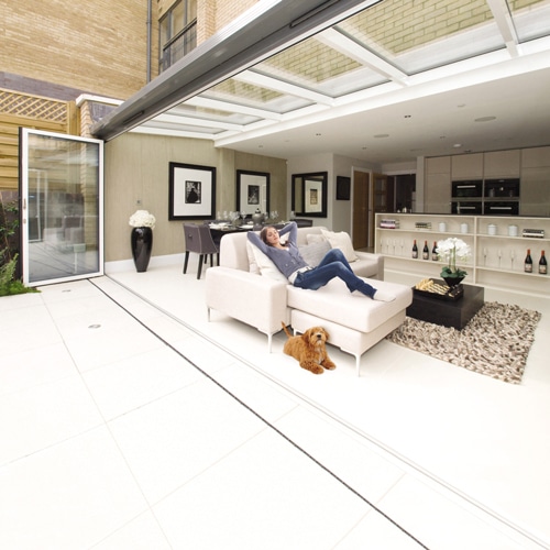 A person relaxes on an indoor chaise lounge in a modern, open-concept living space with a dog lying on the floor. Bifold doors seamlessly blend the interior with the outdoor area.