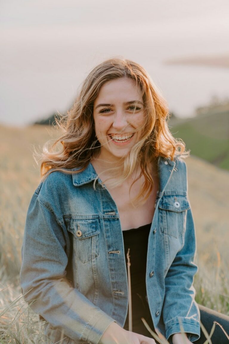 Young woman with long hair wearing a denim jacket sits outdoors in a grassy field, smiling at the camera in natural light.
