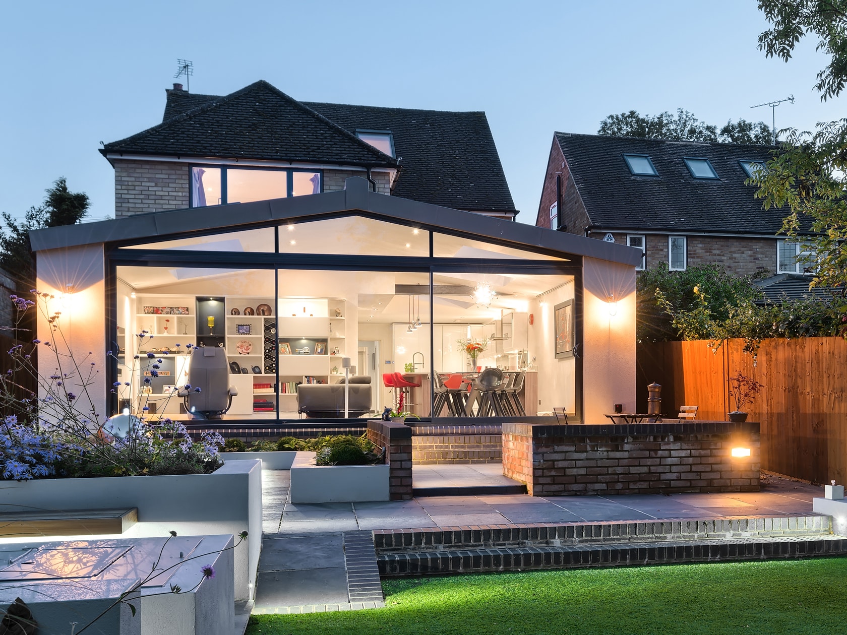 A modern house with a glass-fronted extension showcasing a brightly lit open-plan kitchen, dining, and living space, leading to a landscaped outdoor area.