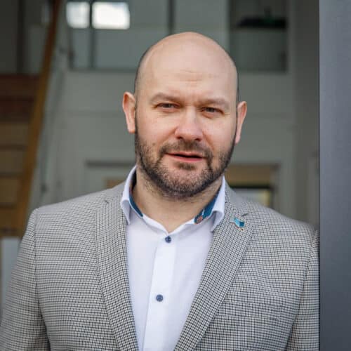 Edward Stobart wearing a light-coloured checked blazer and white shirt, stands indoors near a staircase, looking at the camera.