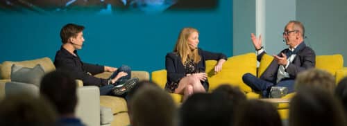 Three people sit on yellow sofas, each wearing a headset microphone, engaged in a discussion in front of an audience.