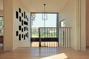 A bright hallway with light wood floors, black railing, wall-mounted shelves, and a large window offering a scenic view of trees and fields.