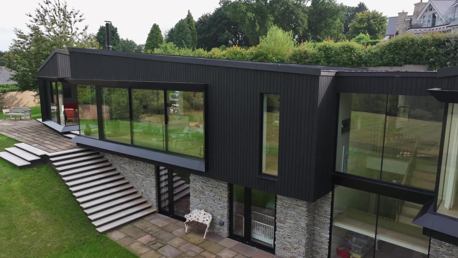 Modern two-story house with large glass windows, black siding, stone walls, and a staircase leading to a patio on a grassy lawn, surrounded by trees.