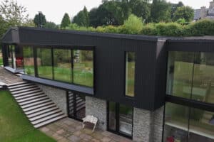 Modern two-story house with large glass windows, black siding, stone walls, and a staircase leading to a patio on a grassy lawn, surrounded by trees.