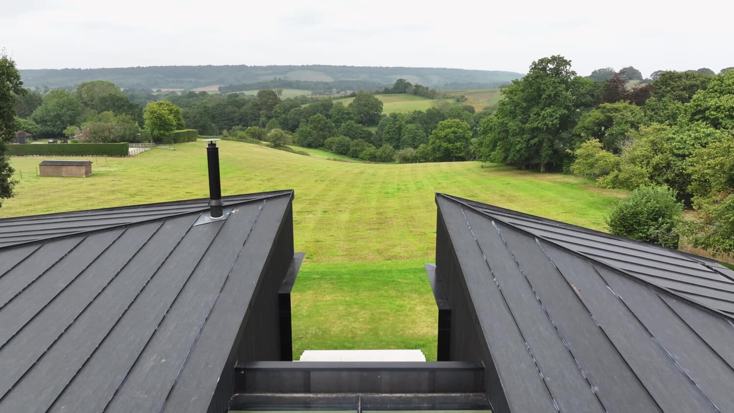 View from a modern black-roofed building overlooking a large, neatly mowed grassy field with trees and hills in the background.