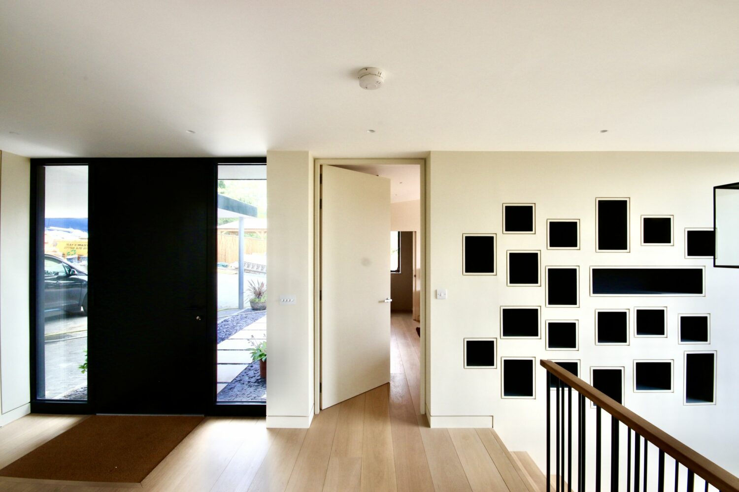 A modern entryway with light wood floors, a large black front door, and a wall featuring multiple empty black picture frames. An open door leads to another room.