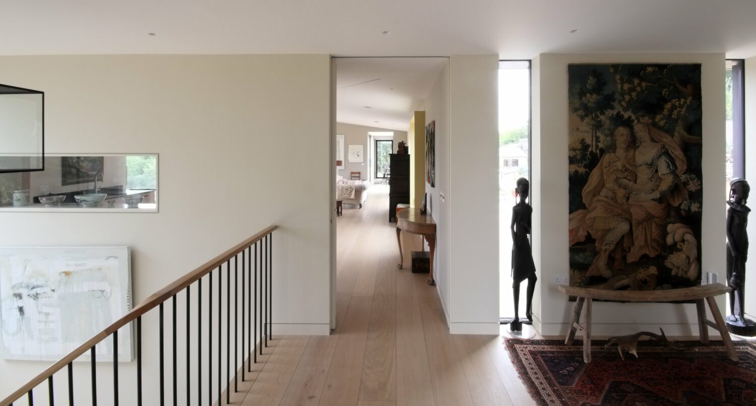 Hallway with light wood flooring, modern railings, artwork on walls, two statues flanking a tapestry, and a view into a bright, open living space.
