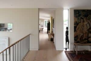 Hallway with light wood flooring, modern railings, artwork on walls, two statues flanking a tapestry, and a view into a bright, open living space.