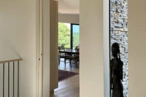 A modern, light-filled interior with minimal furnishings, wooden floors, a dining area with a view of greenery, and a dark sculpture near a stone accent wall.